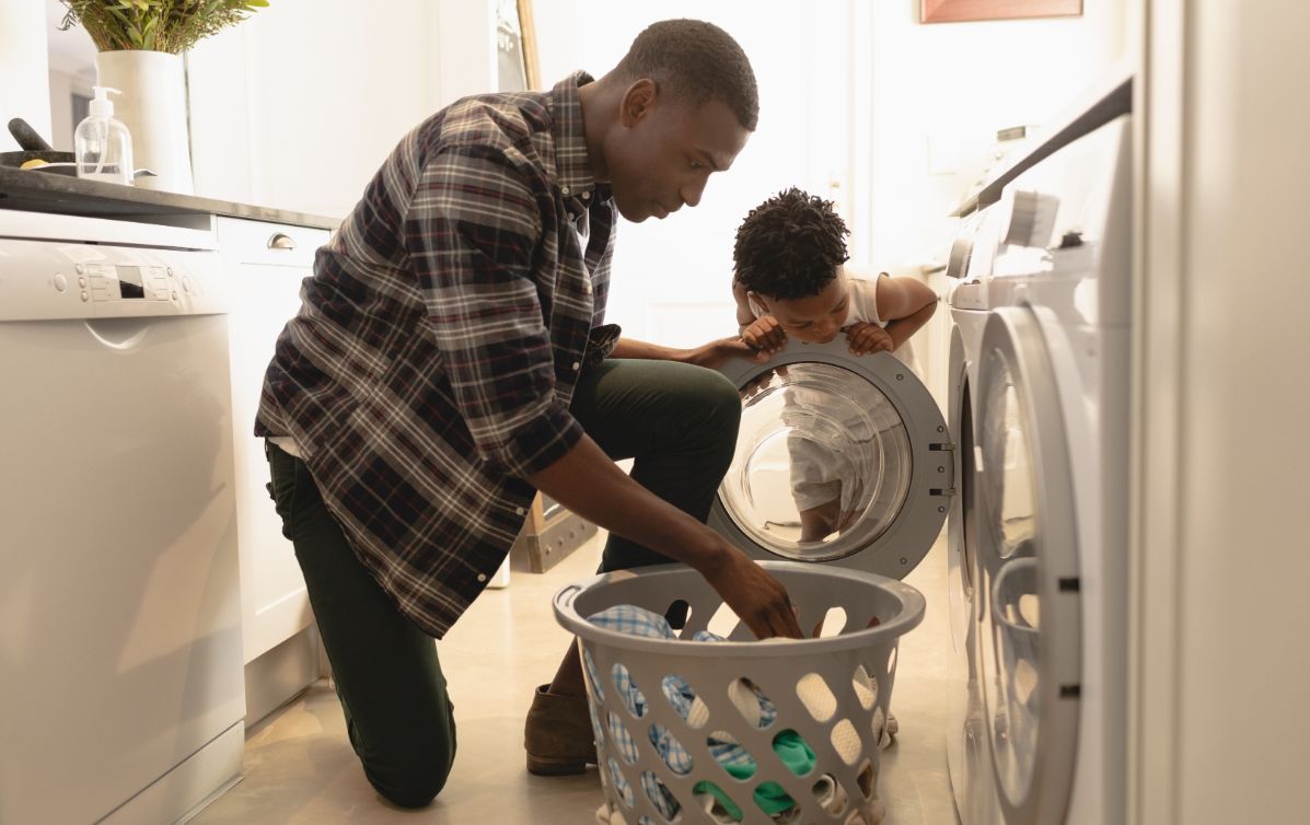 A dad doing the laundry with his boy watching which relates to saving money when using household appliances.